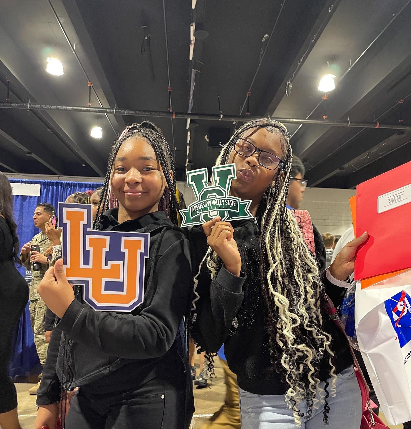 Two young women holding college logos at an educational event, one with a logo for LU and the other with a logo for Mississippi Valley State University.