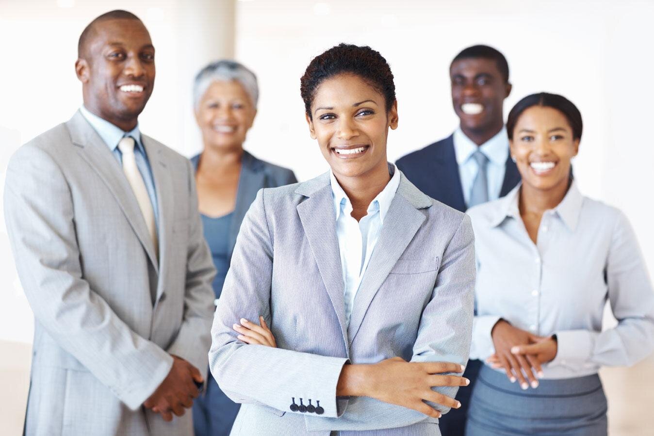 A professional group of five diverse individuals, including a woman in the foreground smiling confidently with her arms crossed, and men and women behind her all dressed in business attire.