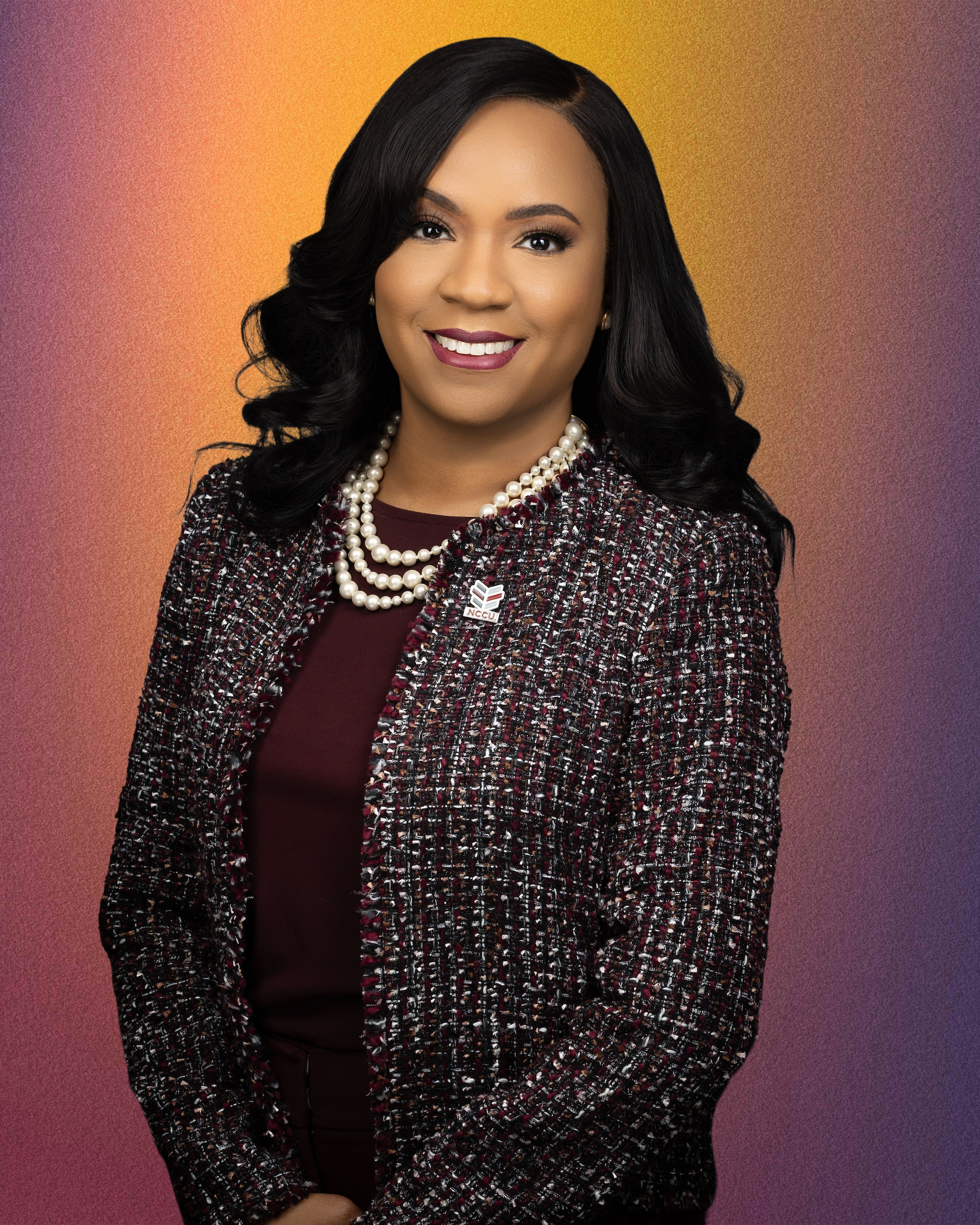 A professional portrait of Dr. Karrie G. Dixon, Chancellor of North Carolina Central University, smiling and wearing a pearl necklace and a patterned blazer, set against a colorful gradient background.