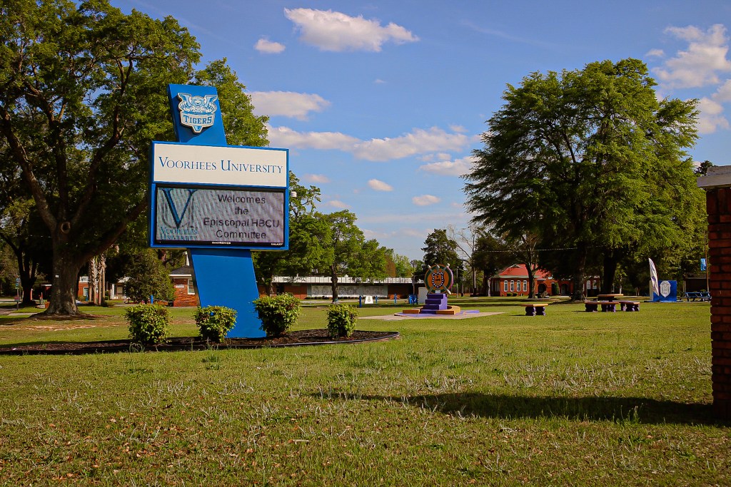 Signage of Voorhees University welcoming the Episcopal HBCU Committee, surrounded by greenery and buildings under a blue sky.