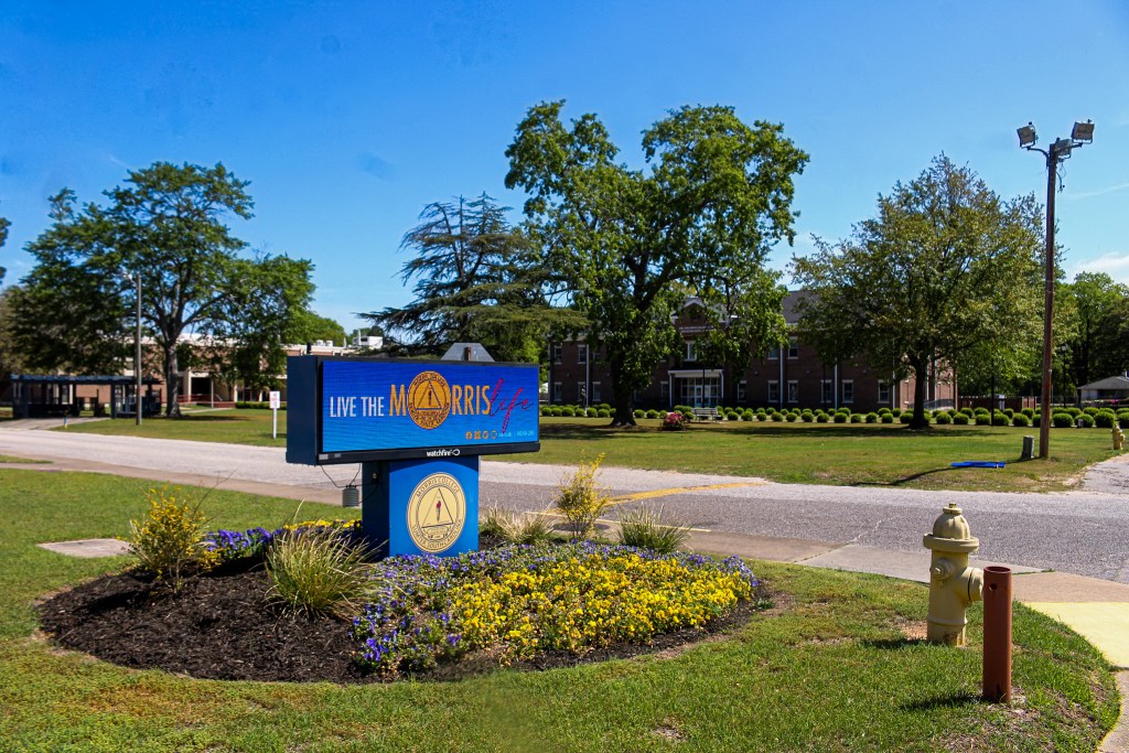 Sign for Morris Life with the text 'LIVE THE MORRIS life' in a landscaped area, featuring trees and flowers, near a brick building.