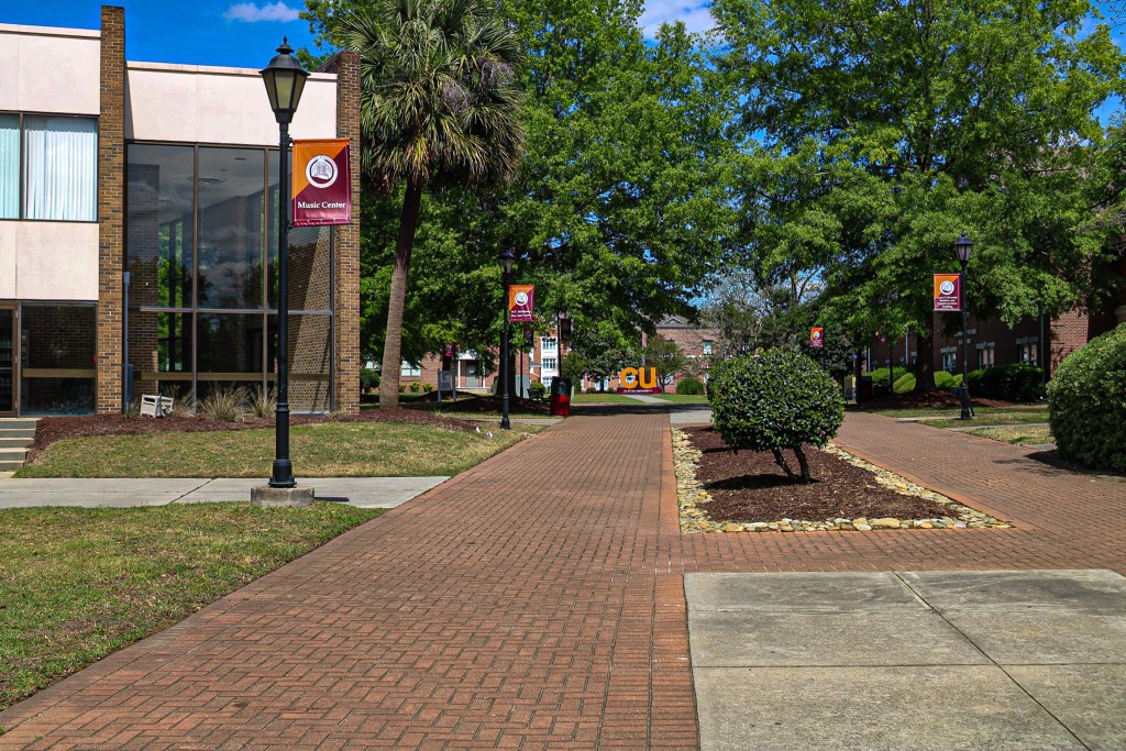 A view of a campus walkway leading to the Music Center building, lined with trees and lampposts. College banners are visible along the pathway.